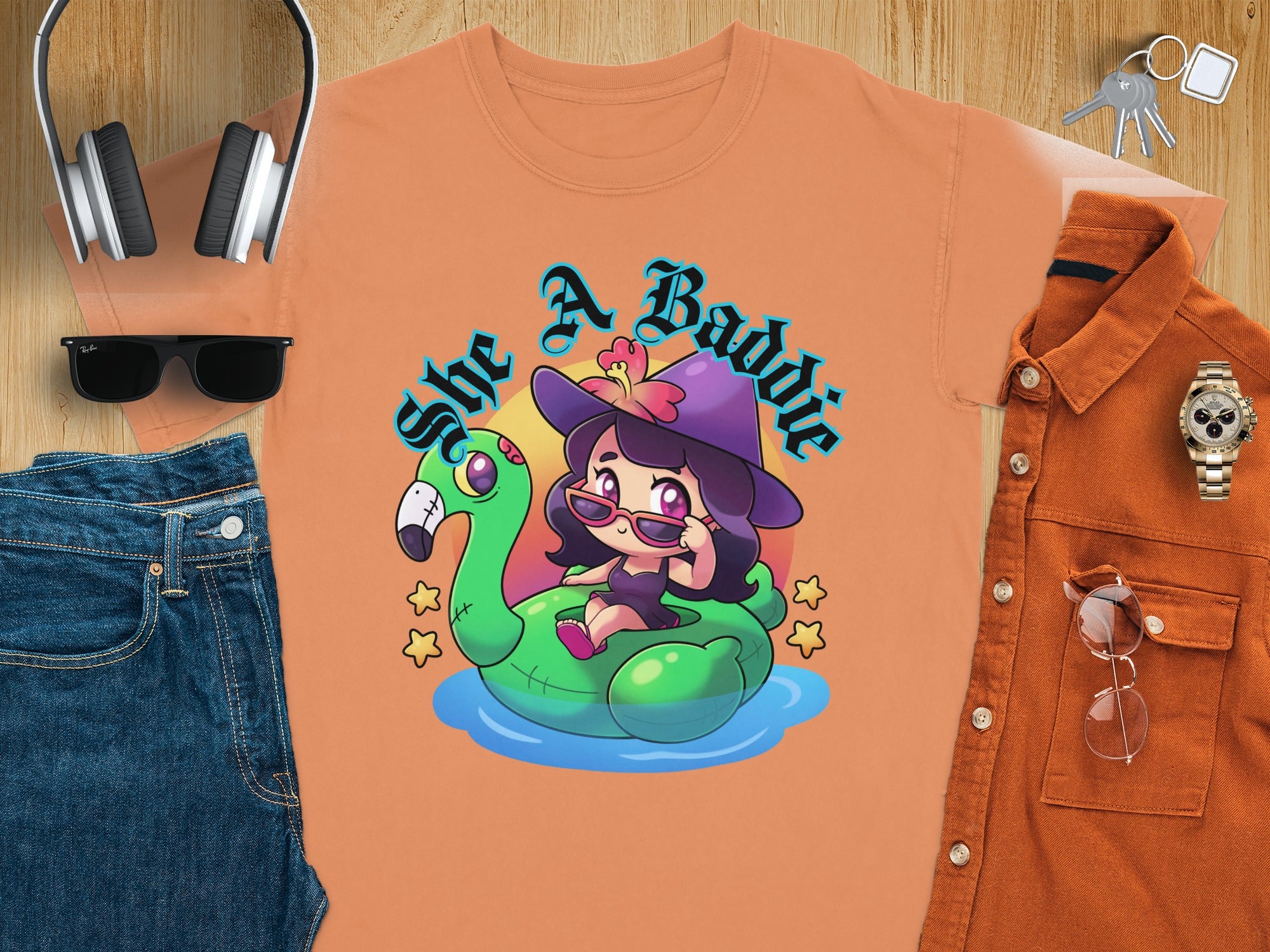 Orange t-shirt with "She A Baddie" text and cartoon witch on flamingo float.