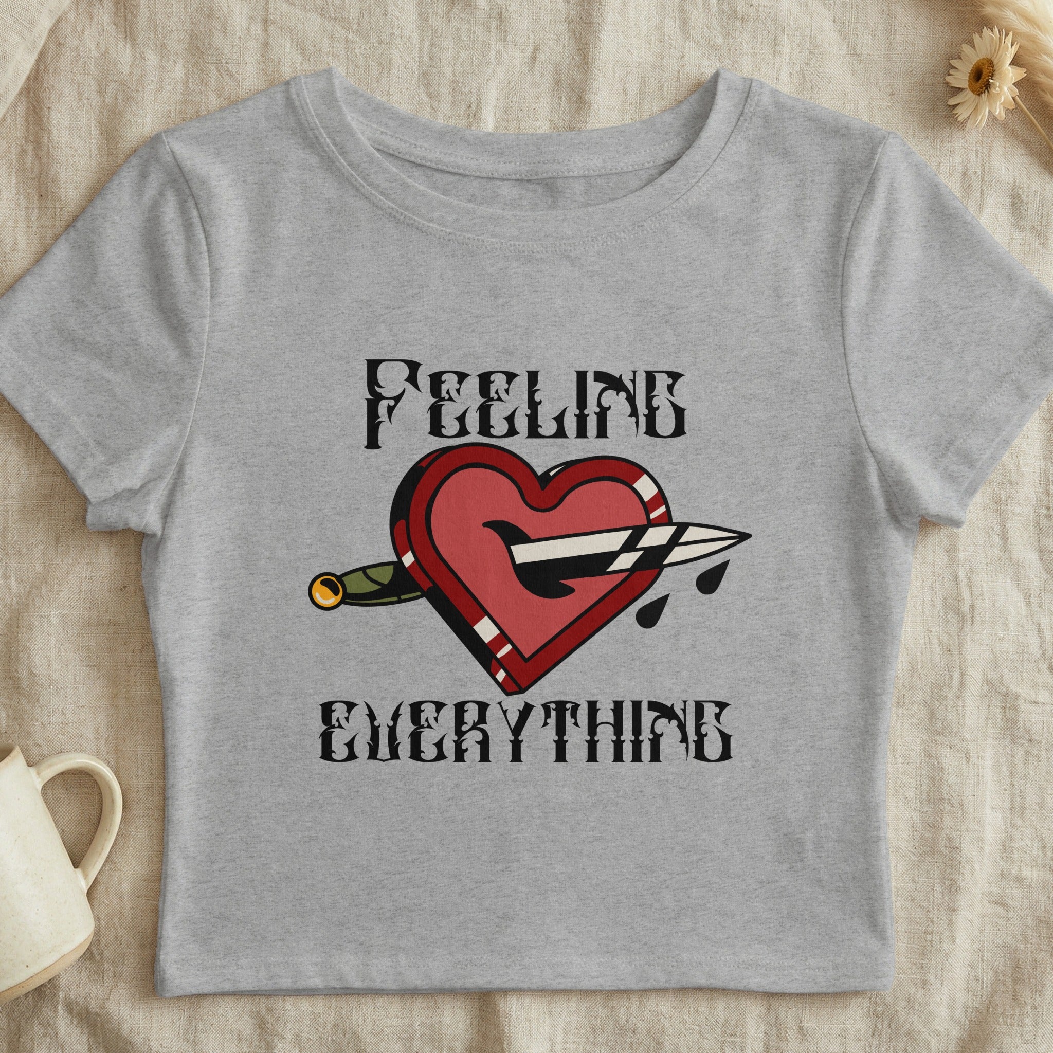 Grey crop top with "FEELING EVERYTHING" text and a heart pierced by a dagger.