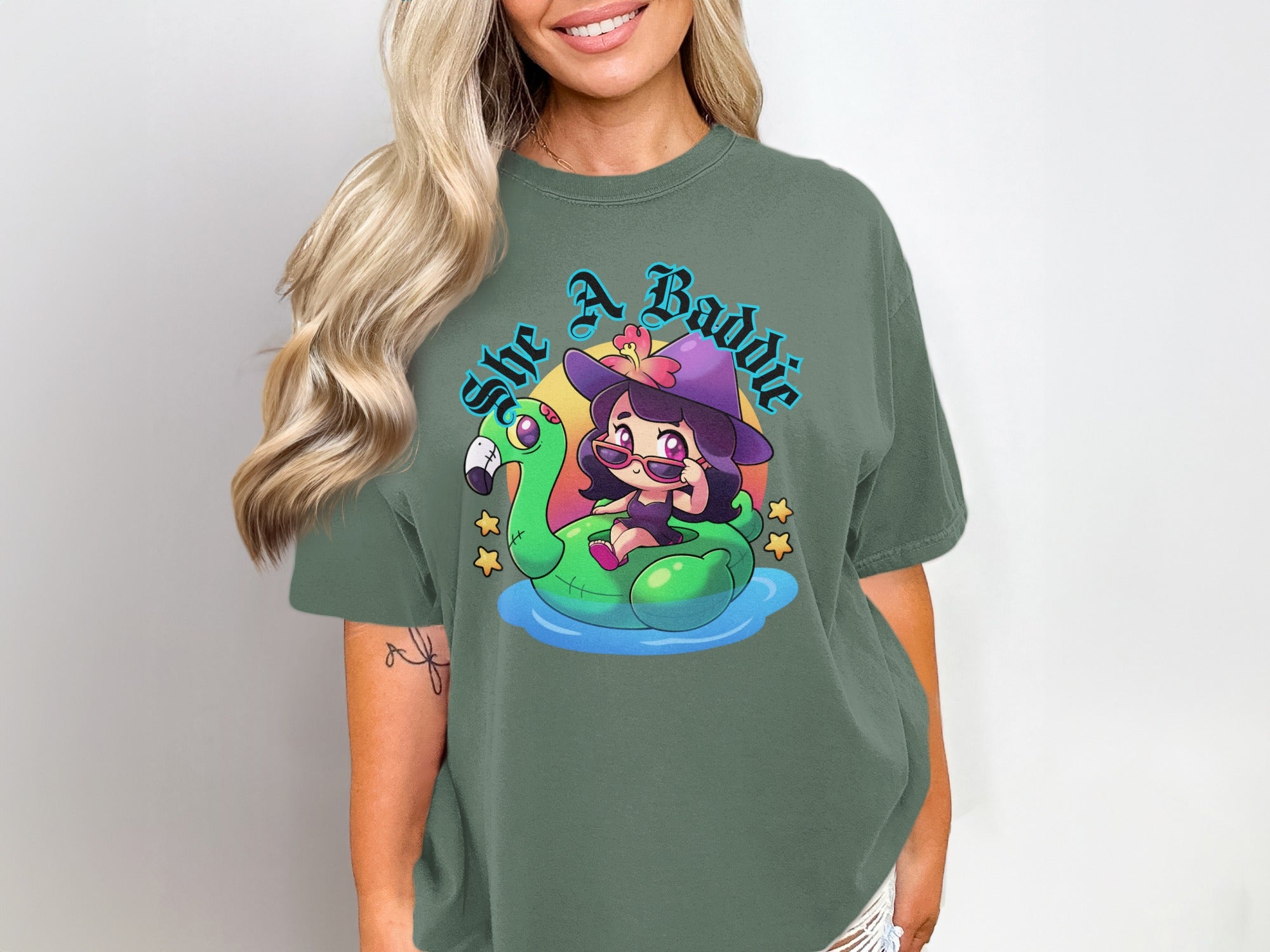 Green t-shirt with a cartoon girl in a witch hat on a flamingo float with text "She A Baddie".