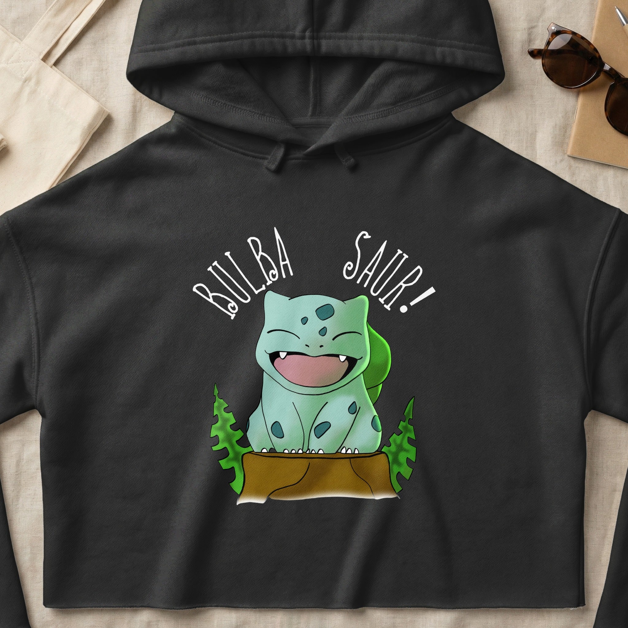 Black hoodie with Bulbasaur character from Pokemon smiling in a woodland scene.