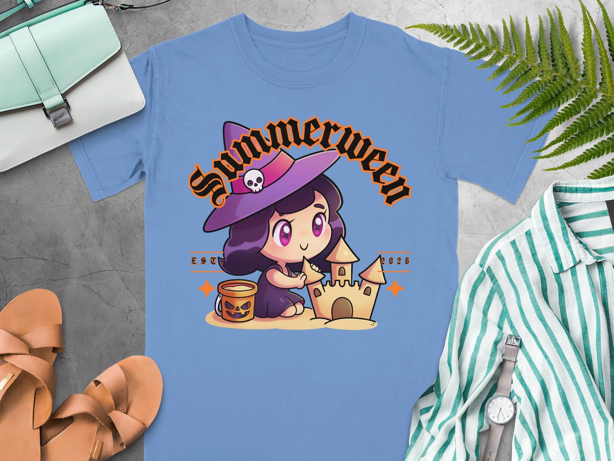 Light blue t-shirt with a cartoon witch building a sandcastle, text "Summerween EST 2026".