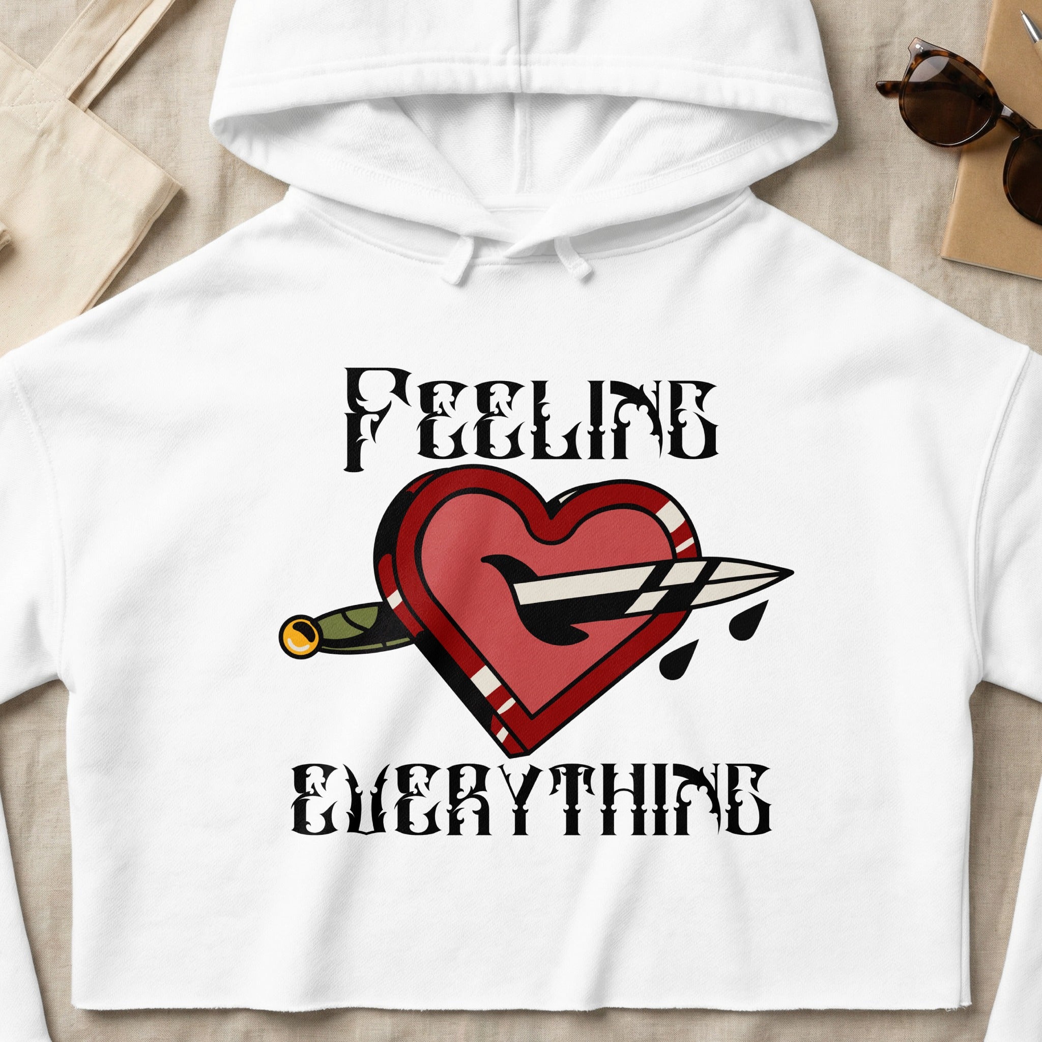 White cropped hoodie with "FEELING EVERYTHING" text and a heart pierced by a dagger design.