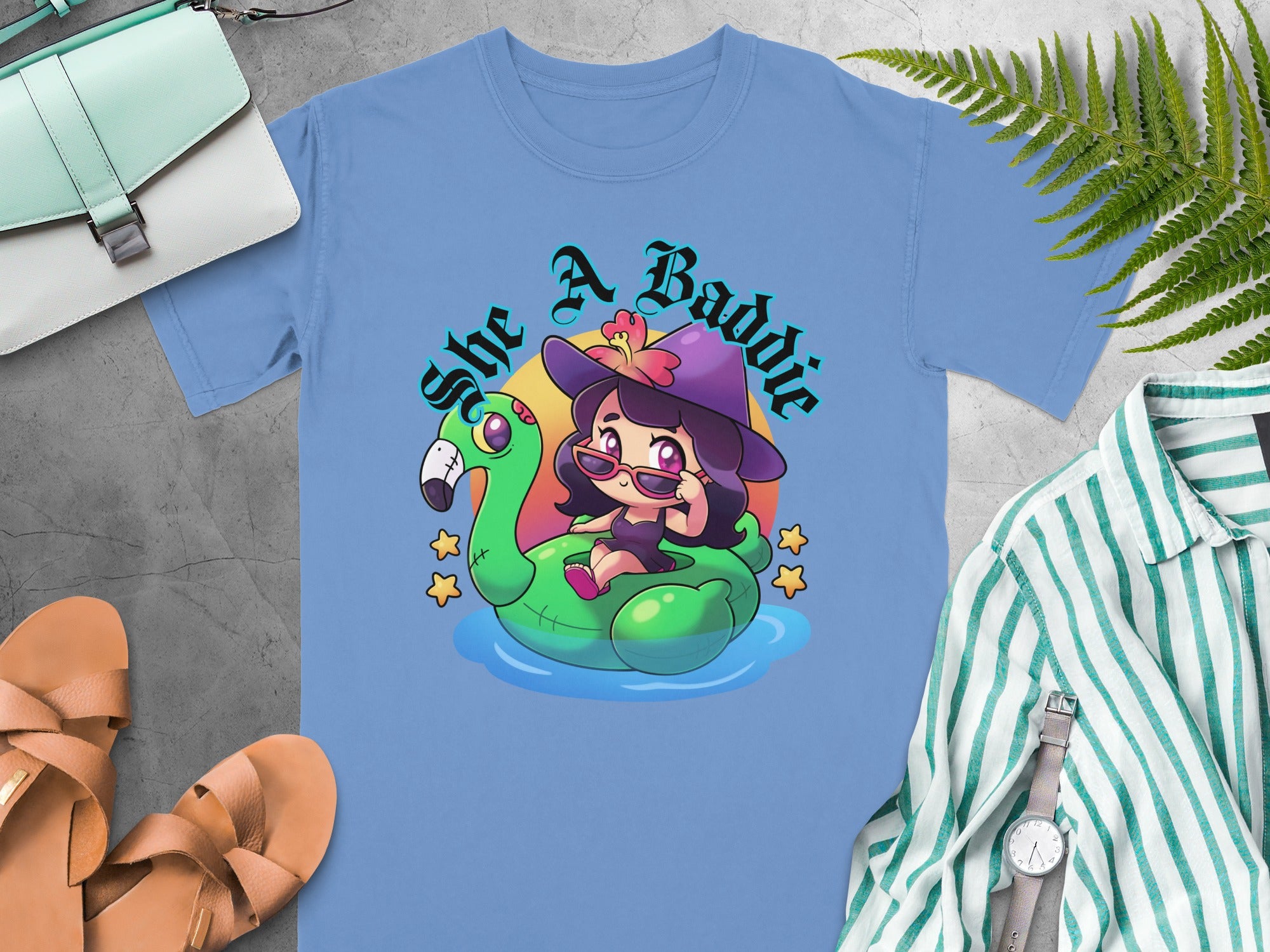 Light blue t-shirt with cartoon of girl in witch hat on flamingo float, "She A Baddie" text.