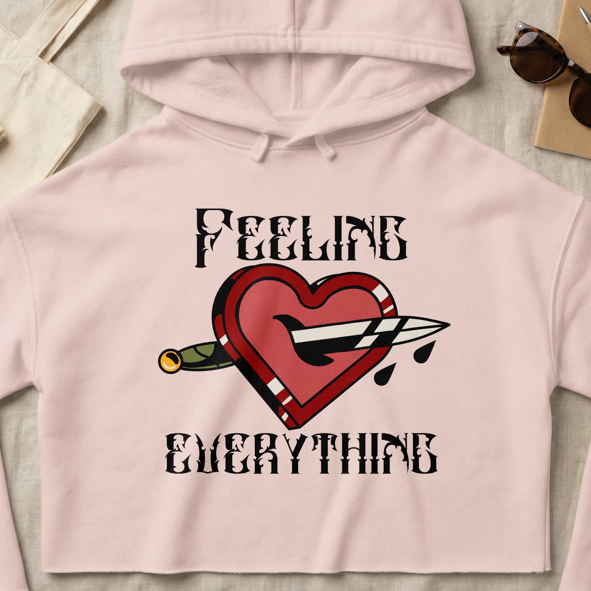 Pink cropped hoodie with heart pierced by dagger and "FEELING EVERYTHING" text.