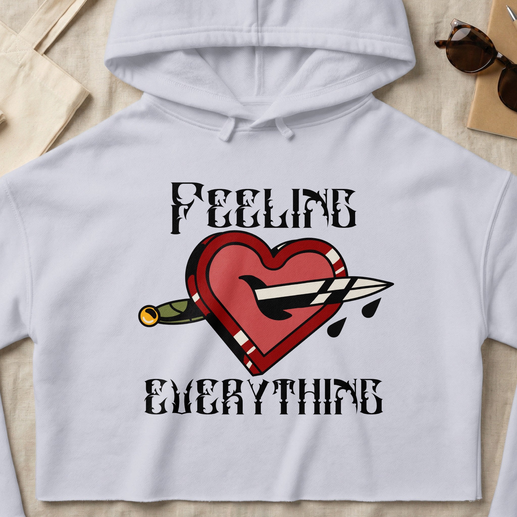 White cropped hoodie with a red heart pierced by a dagger, "FEELING EVERYTHING" text.